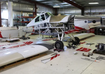 A disassembled white and red agricultural aircraft inside a maintenance hangar, showing the exposed fuselage frame and wings removed for servicing.
