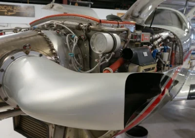 A detailed close-up of a turboprop aircraft engine with its cowling removed, showing the complex network of hoses, wires, and the exhaust ector inside a hangar.